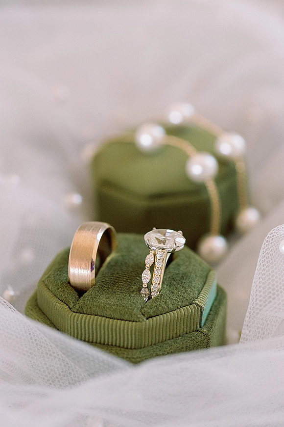 Wedding rings with an engagement ring close up displayed on a ring box beside a pearl strand and tulle on a soft neutral backdrop