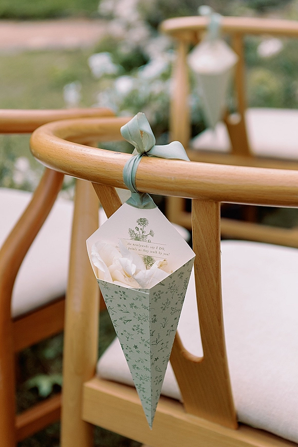 Wedding confetti cones with petal toss cones tied by ribbon bows hang from wooden chairs along a garden lawn ceremony aisle