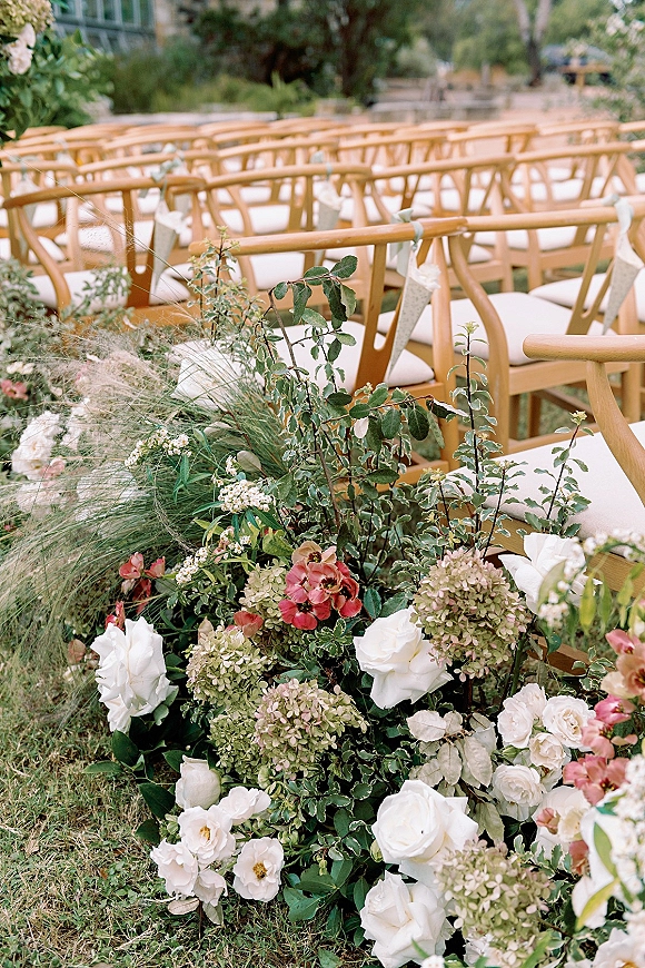 Ceremony aisle decor with wood chairs and low white rose, hydrangea, and pink ground florals lining a garden lawn aisle among trees