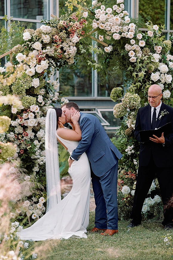 Wedding kiss under a round floral arch of white roses and hydrangeas, bride’s veil flowing beside officiant on a garden lawn