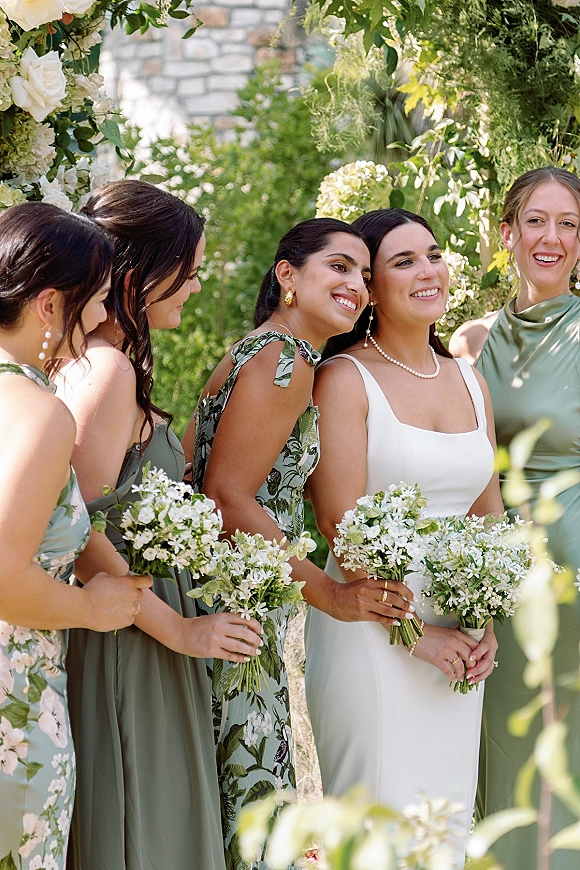 Bridesmaid group photo of bride with bridesmaids in sage green dresses holding white bouquets beneath a floral arch in a garden setting