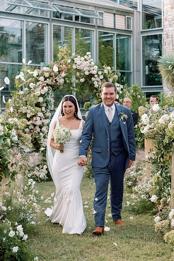 Wedding recessional as bride and groom walk the aisle holding hands, flower petals falling, bouquet and veil under a floral arch on a garden lawn