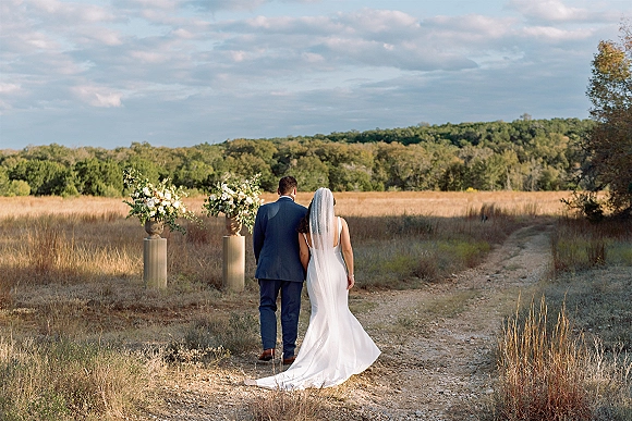 Couple portrait of bride and groom walking away down a dirt road, her long veil trailing behind, in a cloudy meadow field