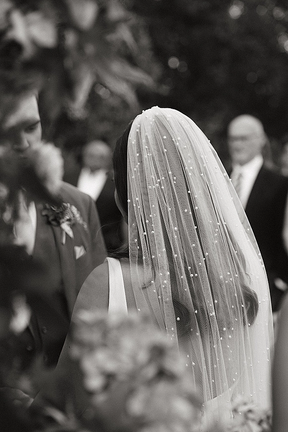 Ceremony moment as bride from behind in a pearl veil faces the groom, holding a bouquet with wedding guests blurred in greenery background