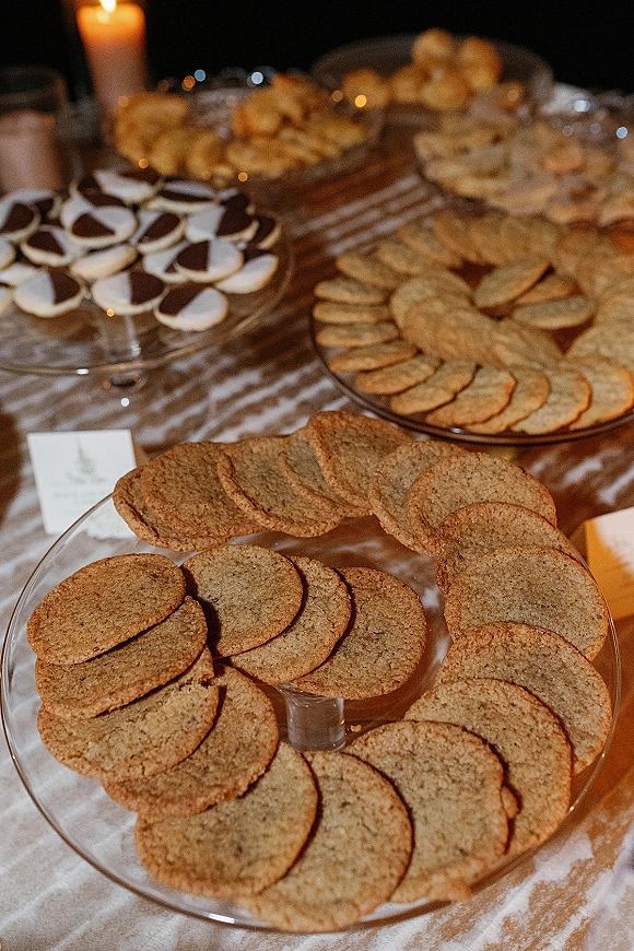 Wedding dessert table with cookie dessert table platters on glass serving trays, place cards and a candle under dim reception lighting