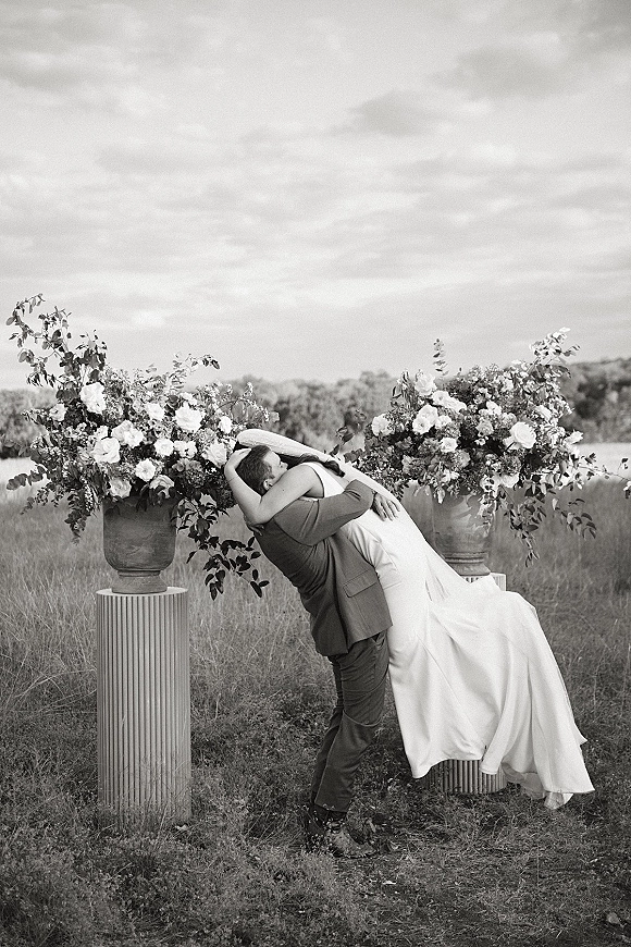 Wedding kiss portrait of bride in a satin gown and groom in a dark suit dipping between floral urns on pedestal columns in a meadow