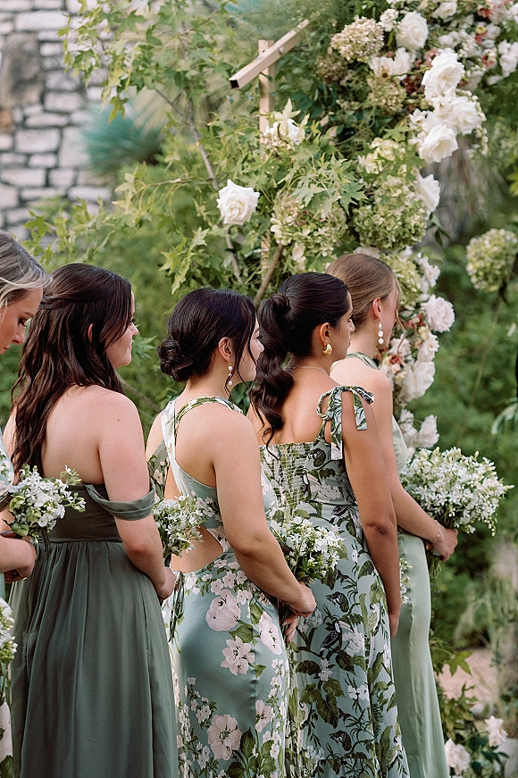 Bridesmaids ceremony lineup in mismatched sage dresses holding white rose and hydrangea bouquets beneath a floral arch by a stone wall