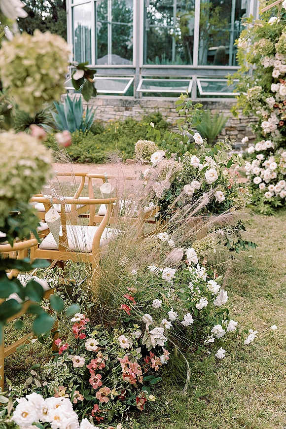 Ceremony aisle flowers with white roses and blush blooms arranged low along a garden lawn aisle beside wooden chairs and greenhouse windows