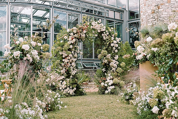 Wedding ceremony arch with white roses and hydrangeas, greenery, and ground florals set before a glass greenhouse and stone wall backdrop