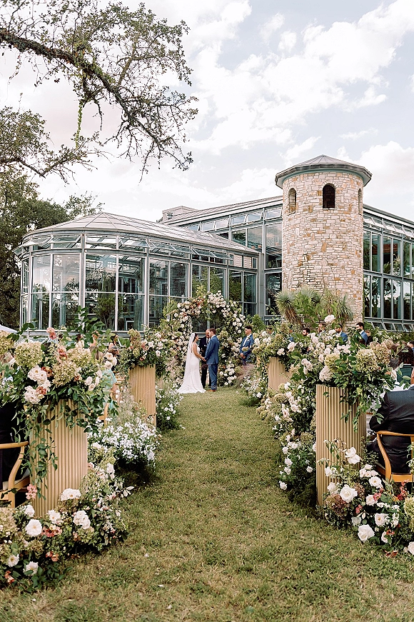 Outdoor wedding ceremony with bride and groom at a flower-lined aisle and floral arch, guests seated by a glass conservatory and stone tower