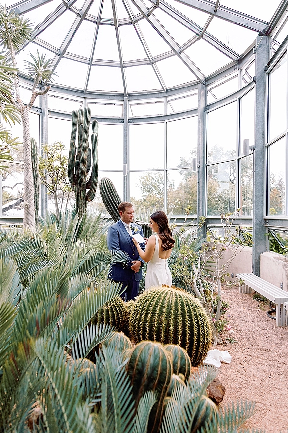 First look moment as bride in a simple satin wedding dress adjusts groom’s boutonniere in a glass greenhouse among cacti on a gravel path