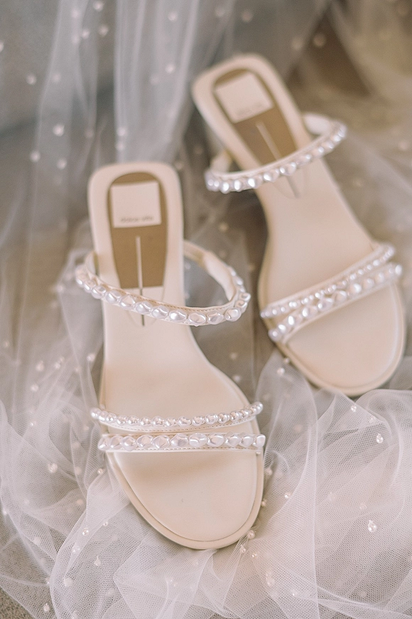 Bridal shoes with pearl wedding shoes straps on a neutral fabric surface, styled with a dotted tulle veil for getting-ready details