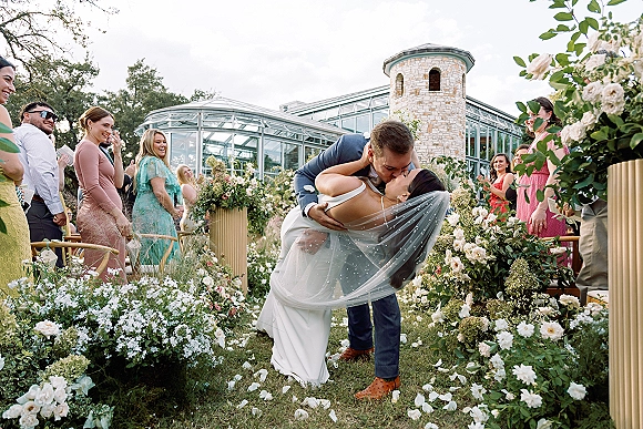 Wedding kiss portrait of bride and groom in a dip kiss, veil trailing, on a flower-lined aisle with guests and a glass conservatory backdrop