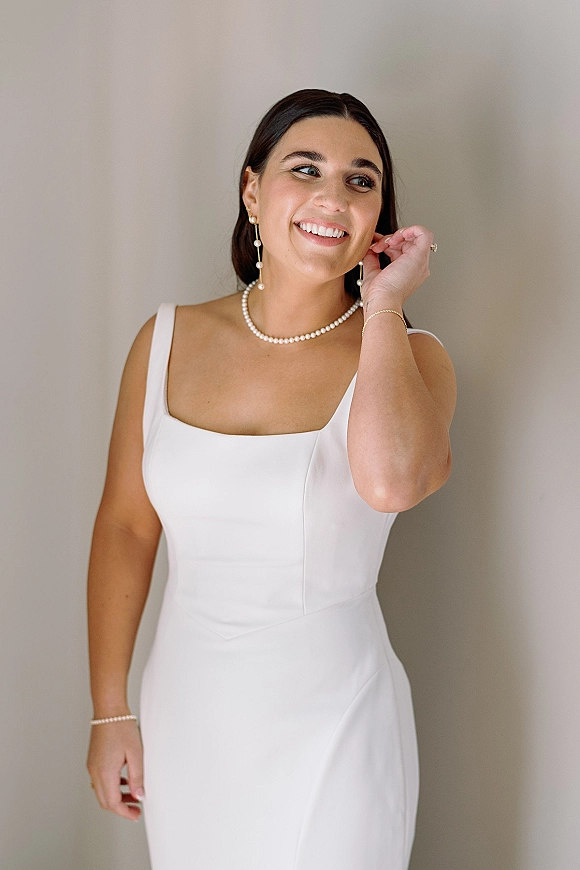 Bridal portrait of a smiling bride in a simple wedding dress, adjusting her pearl drop earring against a neutral wall backdrop