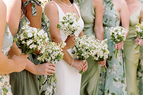 Bridesmaids with bouquets holding small white bridesmaid bouquets in sage green and floral dresses beside the bride in sunlight outdoors