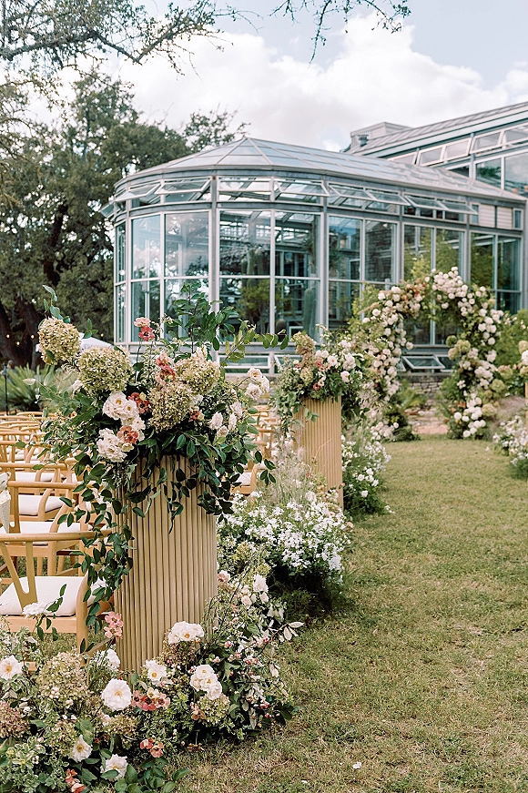 Ceremony aisle decor with hydrangea and garden rose arrangements on pedestal stands, greenery garlands beside wooden chairs in a glass greenhouse