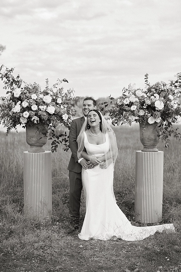 Couple portrait of bride and groom laughing as he hugs her from behind, veil flowing beside floral urns in a grassy field under clouds