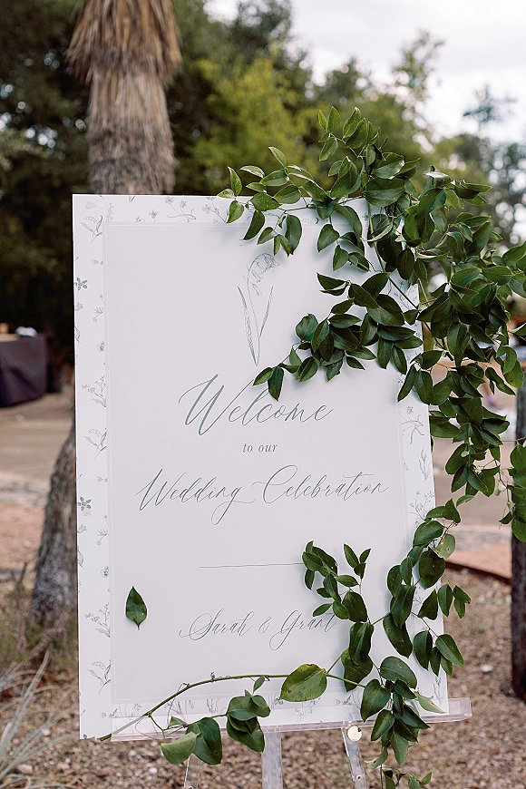 Wedding welcome sign with calligraphy lettering on a clear acrylic stand, framed by greenery garland in an outdoor garden setting