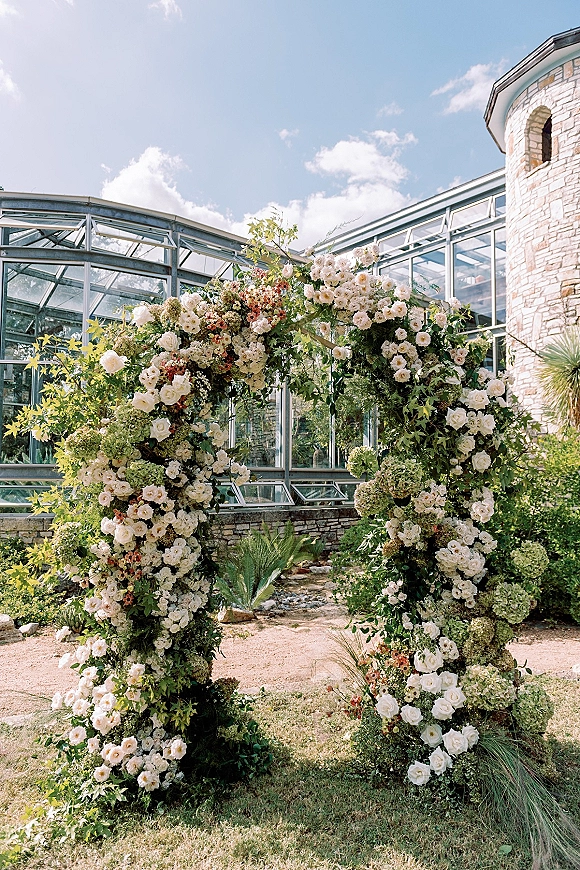 Wedding ceremony arch with a round wedding arch of white roses, hydrangeas, and greenery set before a glass conservatory under blue sky