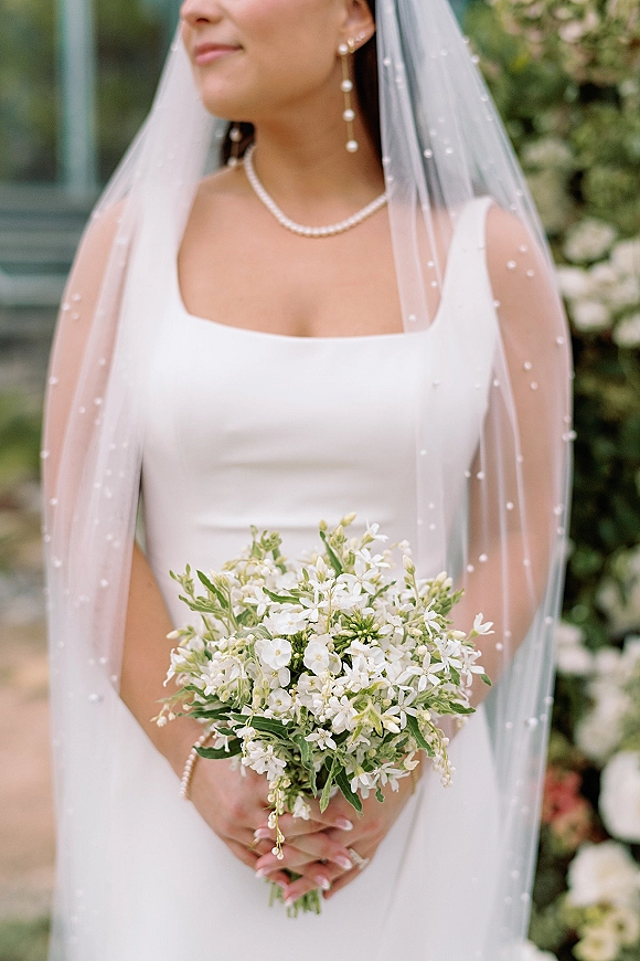 Bridal portrait of a bride holding bouquet in a white dress with pearl veil and jewelry, posing outdoors by greenery and a building exterior