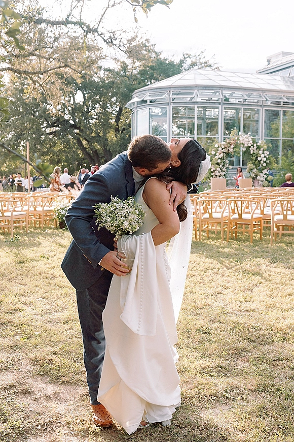 Wedding kiss portrait of bride and groom kissing under a floral arch, her veil and bouquet visible, by a glass conservatory with guests behind