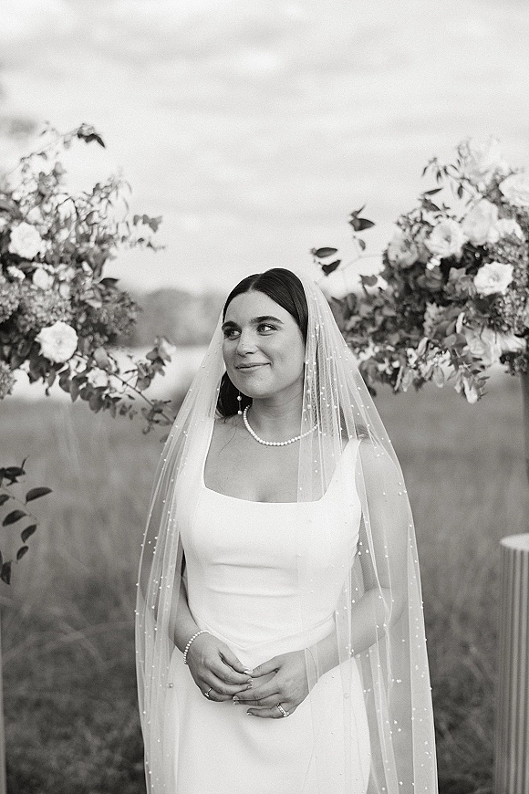 Bridal portrait in black and white of a bride with a long veil over her face, pearl necklace and rings, beneath a floral arch in a field