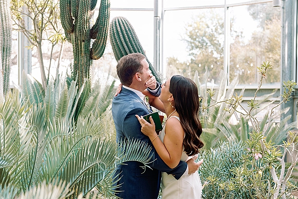 Wedding first look as the bride covers the groom’s eyes, holding a vow book, in a glass greenhouse with cactus and succulents.