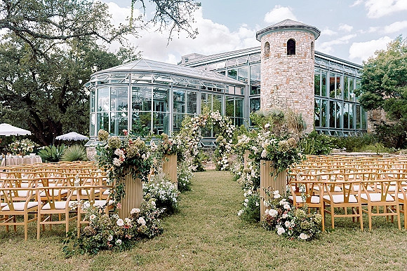 Outdoor ceremony setup with garden wedding ceremony florals lining the aisle, floral arch, and chairs on a lawn by a glass conservatory