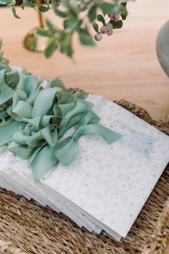 Wedding invitation suite with sage green wedding invitations tied in ribbon bows on a rattan tray, styled with greenery on wood tabletop