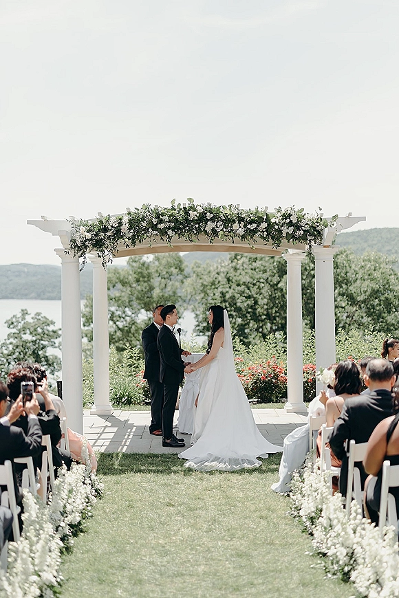 Wedding ceremony moment as bride and groom hold hands at a floral garland arch, with guests seated by a lake and mountains