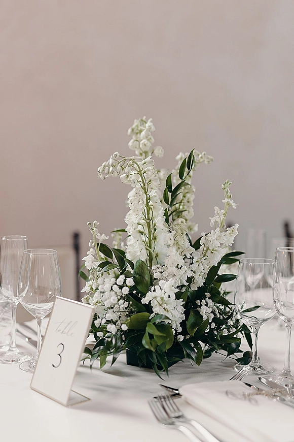 Wedding centerpiece with a white floral centerpiece and greenery, baby's breath, and table number card on a white tablecloth table setting