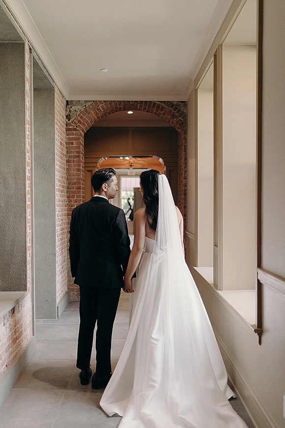 Couple portrait of bride and groom holding hands walking away, her cathedral veil trailing in a brick-arch hallway with windows