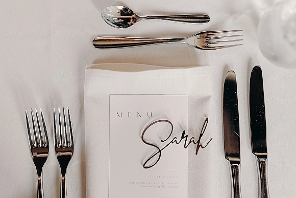 Wedding place setting with a wedding menu card and calligraphy place card on a folded napkin, silver flatware and glassware on white tablecloth