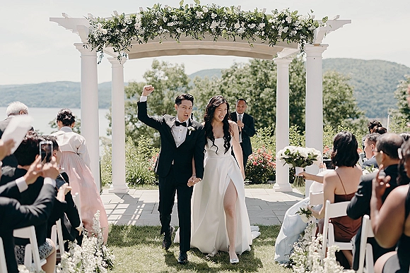 Wedding recessional as bride and groom walk the aisle, cheering past white folding chairs under a pergola with floral garland, mountains and lake beyond