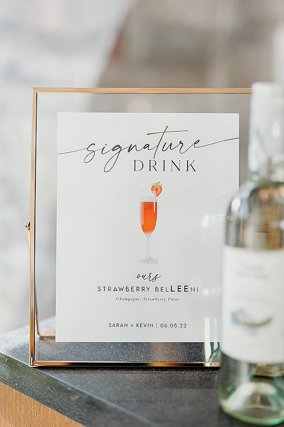 Signature drink sign in a gold frame showing a wedding signature drink menu with cocktail illustration beside a champagne bottle on a bar counter