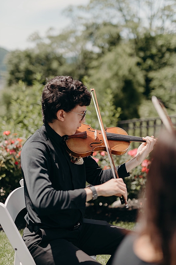 Wedding ceremony musician playing violin with a bow, wearing a black suit and sunglasses, seated by red flowers in a garden setting