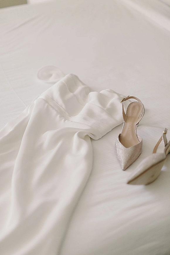 Wedding dress details with pointed toe bridal heels on a white bed, showcasing ankle strap shoes laid neatly on the fabric