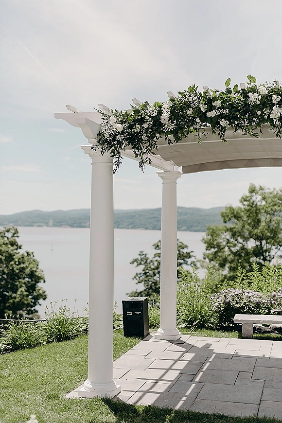 Wedding ceremony arch with a white pergola wedding arch draped in greenery garland and white flowers on a stone patio by a lake