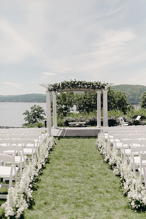 Ceremony setup for an outdoor wedding ceremony with a white pergola, floral garland, and aisle flowers on a stone patio by a lake and hills