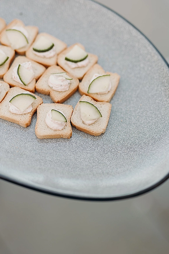 Wedding appetizers with crostini and cucumber slices topped with creamy spread, arranged on a serving plate over a gray tabletop