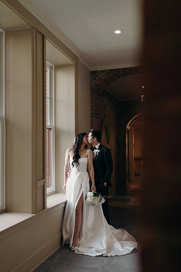 Wedding kiss portrait of bride and groom kissing, her strapless slit gown and bouquet in window-lit hallway with brick archway