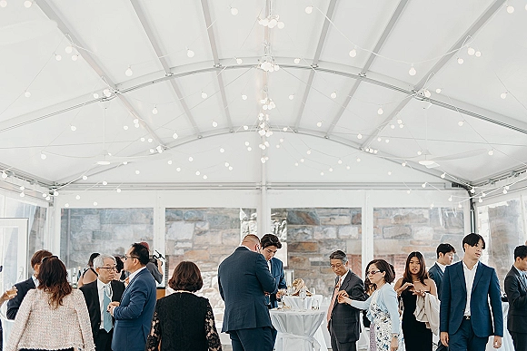 Wedding cocktail hour with wedding guests mingling around high top tables under a white tent with bistro lights and wine glasses