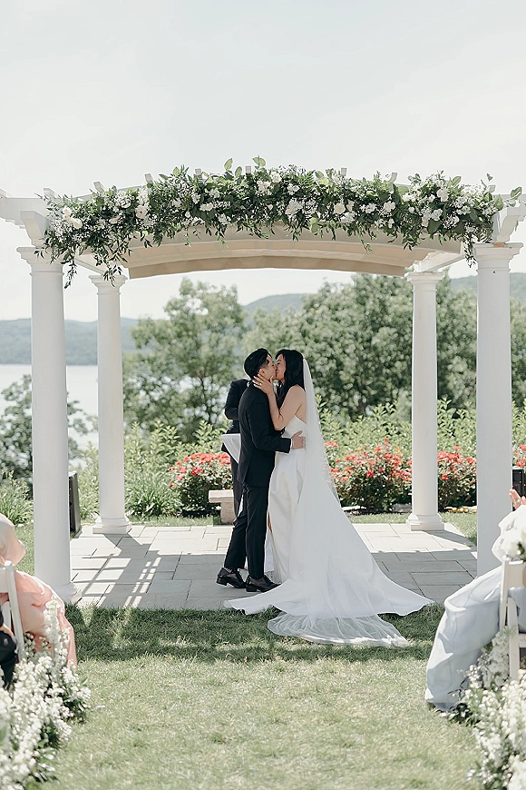 Wedding kiss beneath a white-column arch with greenery garland, bride in strapless dress and veil, lake and hills behind