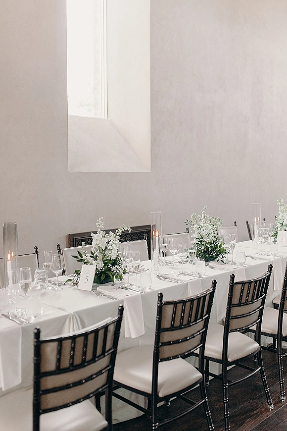 Reception tablescape with white wedding table decor, white floral centerpieces, glass cylinder candles and taper candles by tall window