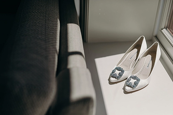 Bridal shoes in white satin pumps with blue crystal buckles on a windowsill, lit by soft window light and casting shadows