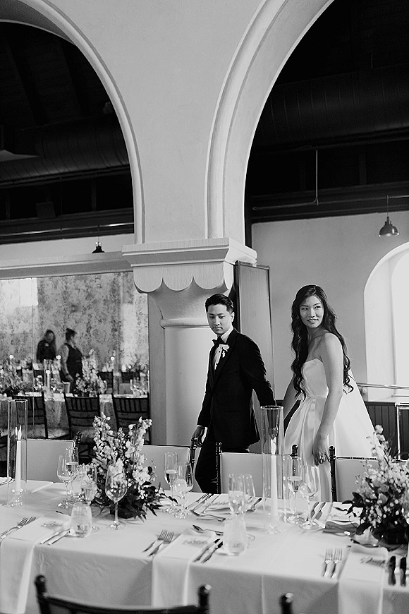 Wedding reception entrance as newlyweds enter, bride in strapless gown and groom in tuxedo passing tables with candlelit floral centerpieces under an archway