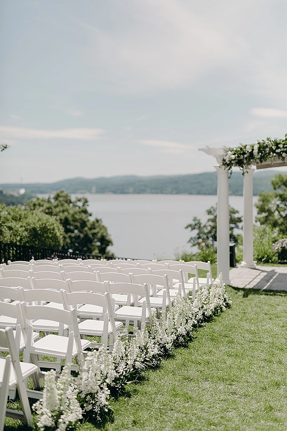 Ceremony setup with white folding chairs lining an aisle of flowers toward a pergola altar on a lakefront lawn under blue sky