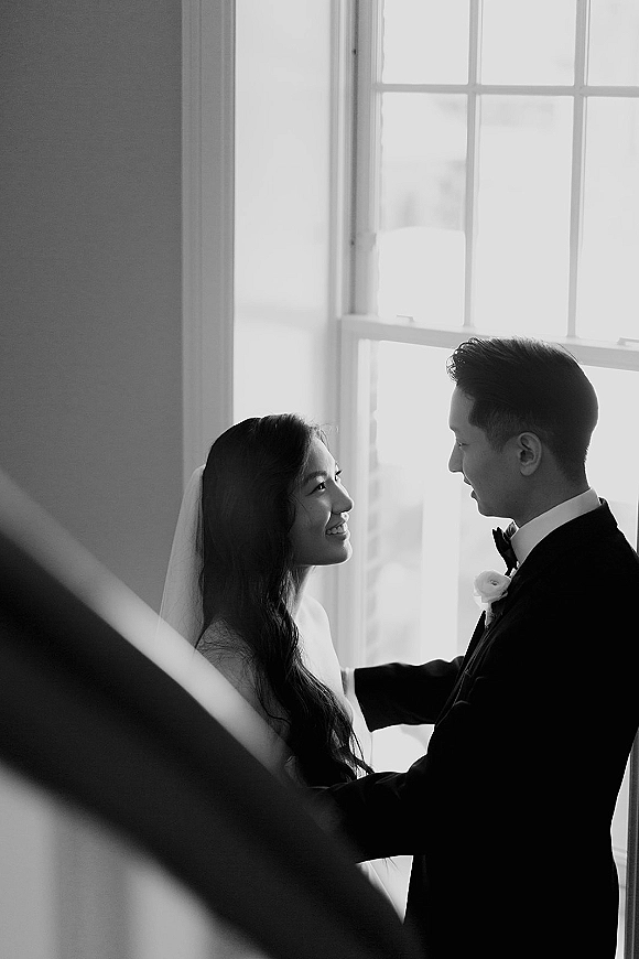 Couple portrait of bride and groom in window light, groom holding her as she smiles, veil and tuxedo by a staircase railing indoors