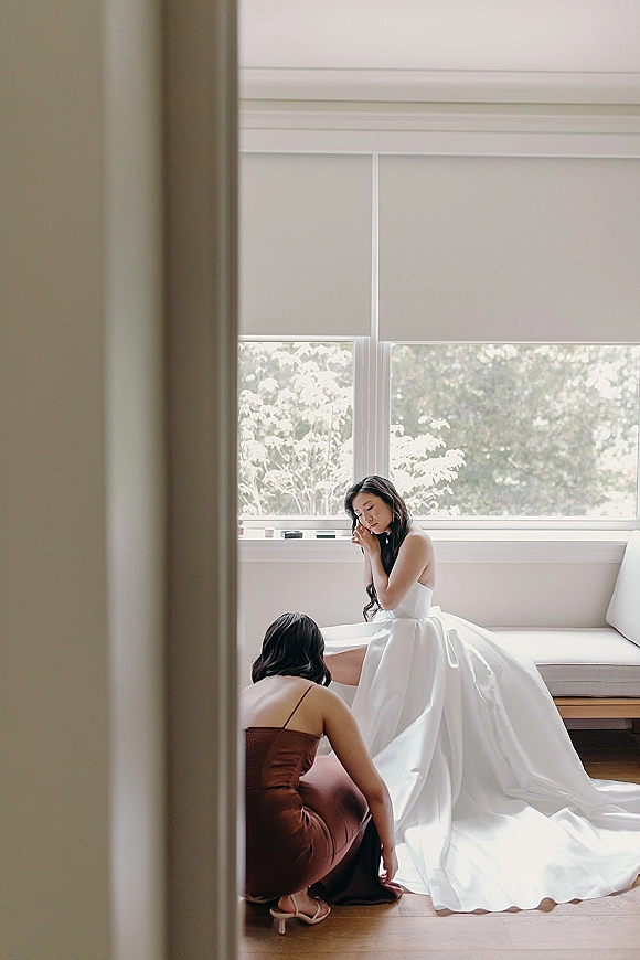 Bride getting ready as a bridesmaid helps bustle her strapless ball gown by a large window, long wavy hair and heels on hardwood floor