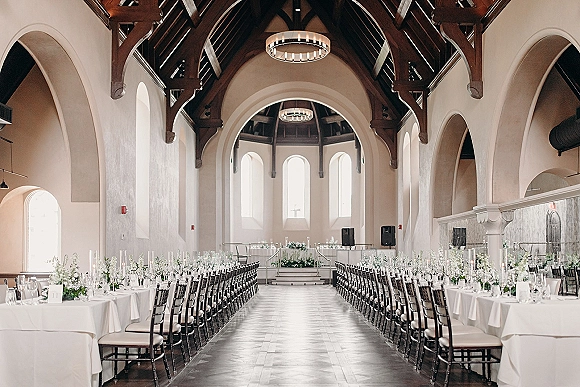 Ceremony setup in a chapel wedding ceremony with a white aisle runner between long banquet tables lit by taper candles under arched windows